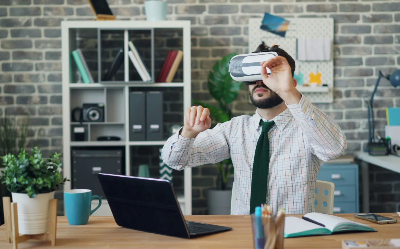 Man wearing a tie and using a virtual reality headset while sitting at a desk with a laptop and office supplies.
