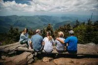 Group of five people sitting on rocks at a mountain overlook, enjoying a scenic view of forested hills under a partly cloudy sky.