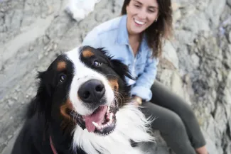 Close-up of a happy dog with a woman smiling in the background while sitting on rocky terrain outdoors.