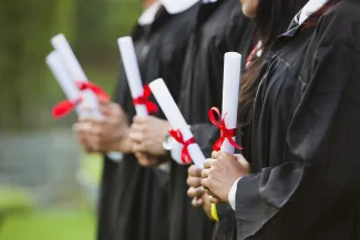 Graduates in black caps and gowns holding rolled diplomas tied with red ribbons during a graduation ceremony.