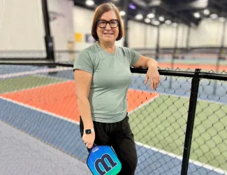 Woman standing by an indoor pickleball court, smiling and holding a paddle while leaning against a chain-link fence.