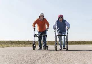Two elderly men wearing helmets racing with walkers on an open road, smiling and appearing playful under a clear sky.
