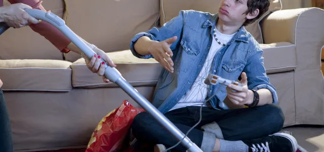 Young man sitting on the floor playing video games, reluctantly handed a vacuum cleaner hose by another person, with a bag of chips beside him.