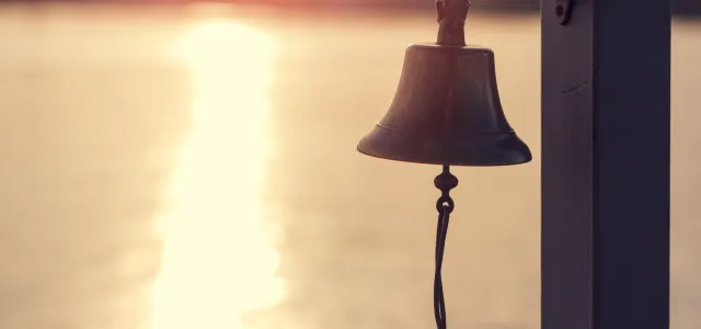 A metal bell mounted on a wooden post, overlooking calm water at sunset with golden light reflecting off the surface.