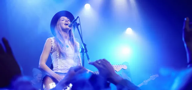 Female musician wearing a hat performing on stage with a guitar and singing into a microphone under bright blue concert lights, with an audience cheering in the foreground.