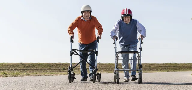 Two elderly men wearing helmets racing with walkers on an open road, smiling and appearing playful under a clear sky.